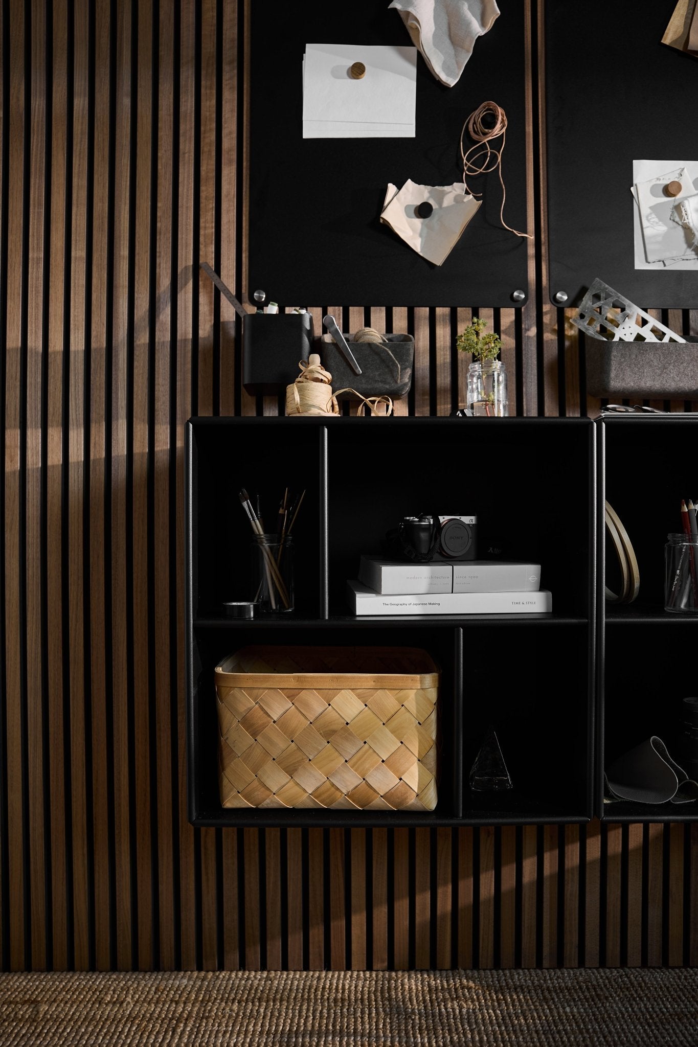 The Arka Open Cabinet in Black by WoodUpp beneath a black bulletin board, books, camera, decor, and woven basket against a Nordic acoustic slatwall panel.